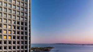 Un edificio alto con una fachada de ventanas que da a una masa de agua tranquila con barcos y una costa lejana.