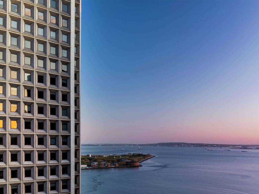 Un immeuble de grande hauteur, doté d’une façade à fenêtres disposées en réseau, surplombe un plan d’eau paisible où l’on aperçoit des bateaux et une côte lointaine.