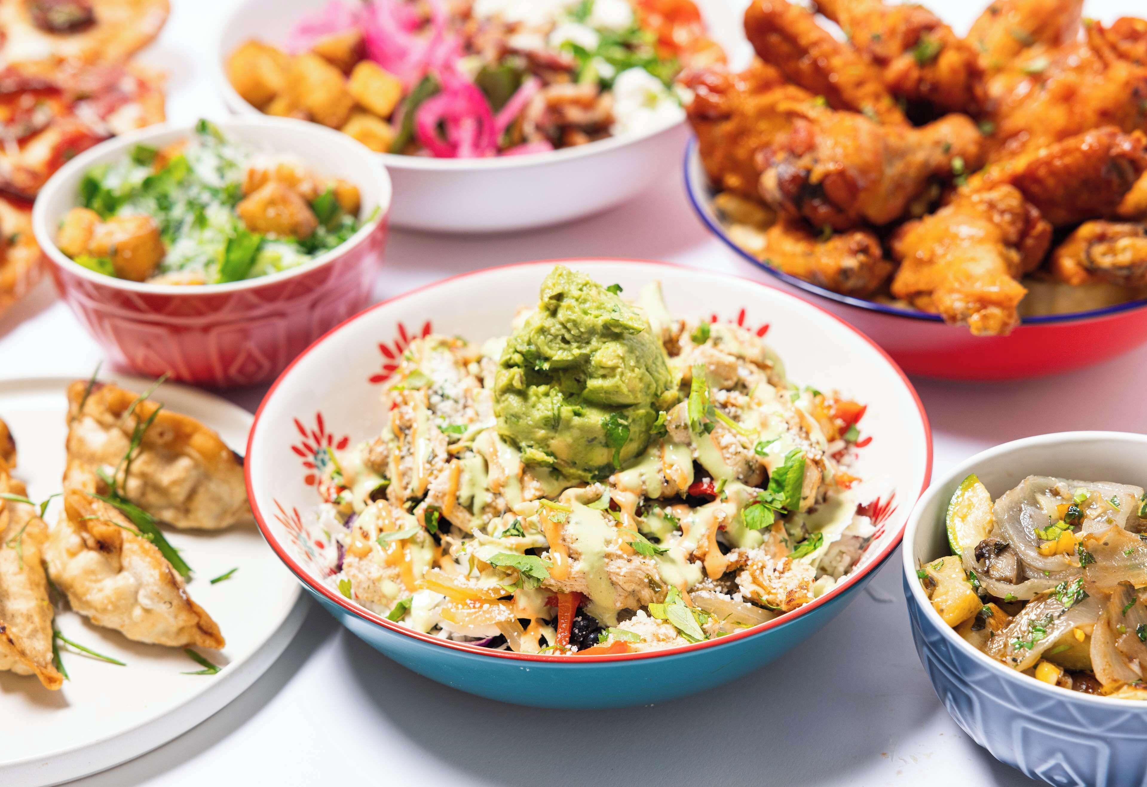 Una mesa con diversos platos, incluyendo un tazón de ensalada de aguacate, alitas de pollo fritas y otras preparaciones variadas.