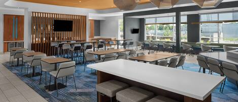 A modern hotel lobby with a central bar area, multiple seating arrangements, and large windows offering views of the outdoors.