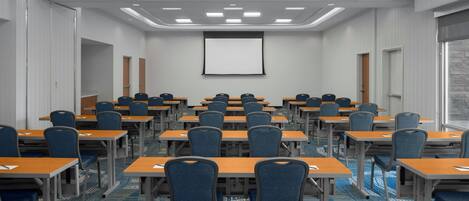 A conference room with rows of tables and chairs, a projector screen, and a carpeted floor.