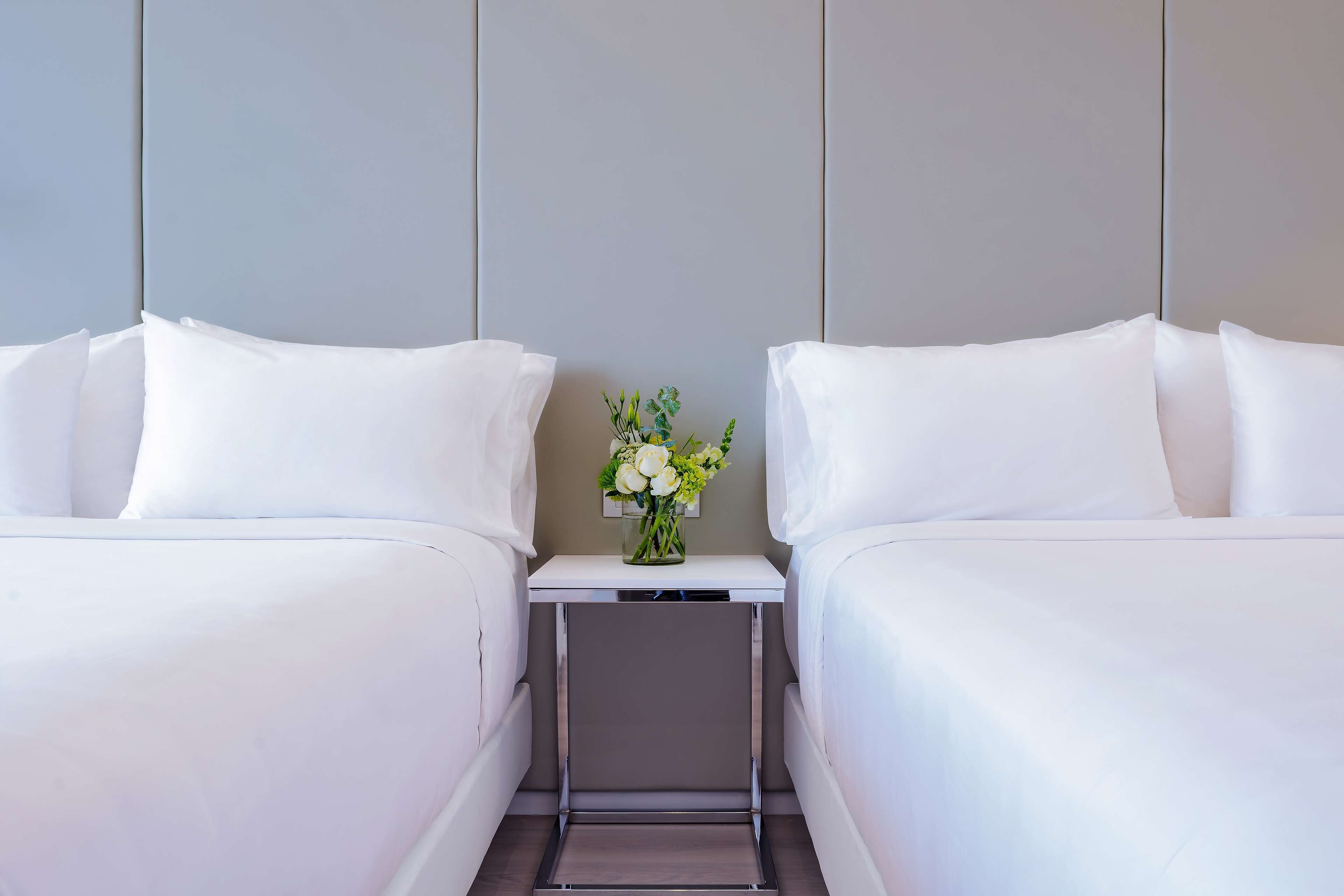 Habitación de hotel con dos camas individuales, una mesita de noche con un florero y un cabecero gris.