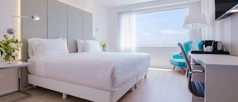 A modern hotel room with a large bed, a desk with a chair, a TV, and a window with a view of the sky.