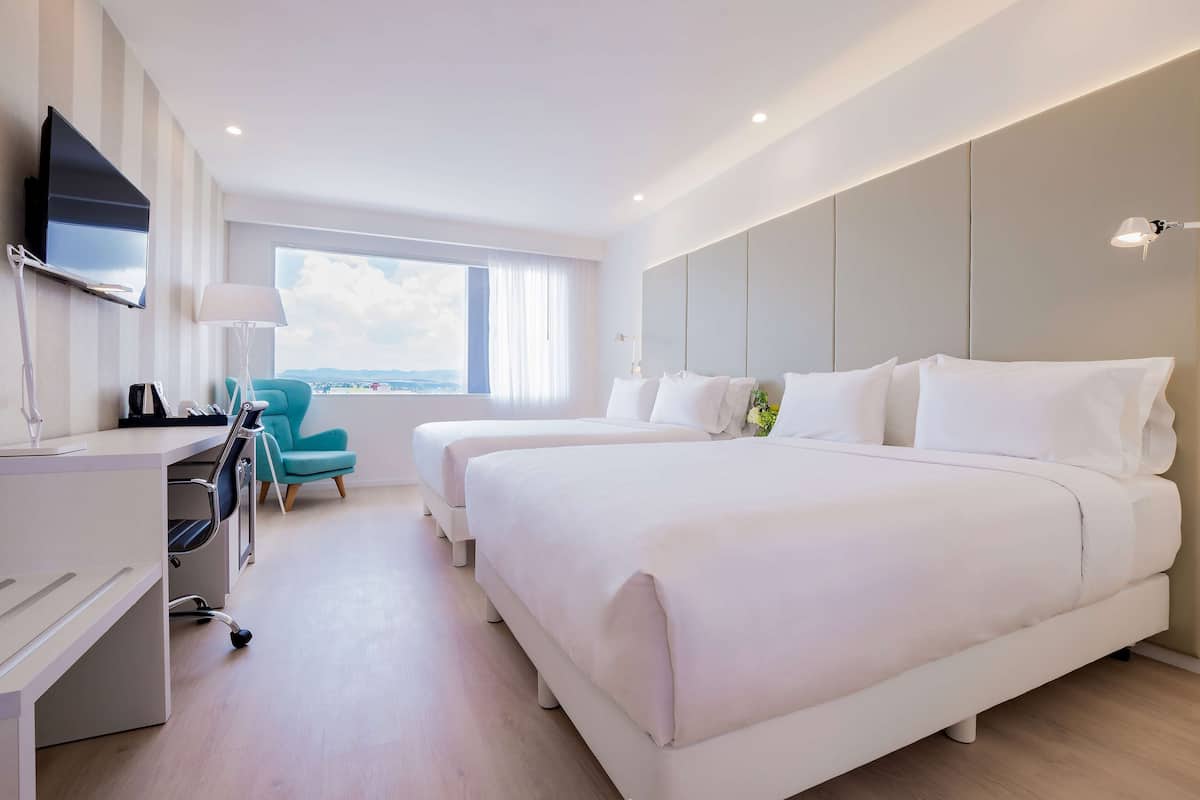 A modern hotel room with a large bed, a desk with a chair, a TV mounted on the wall, and a window with a view.