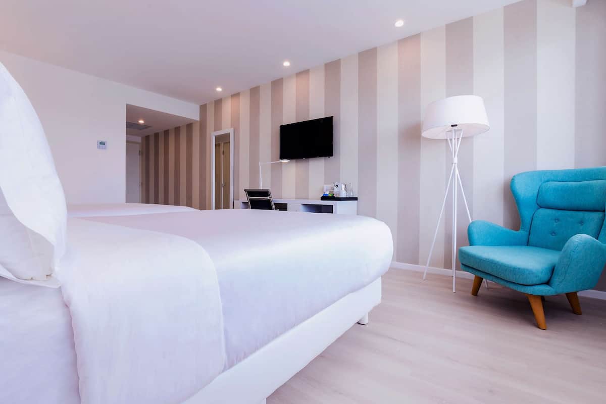 A modern hotel room with a large bed, a desk with a computer, a flat-screen TV, and a blue armchair.