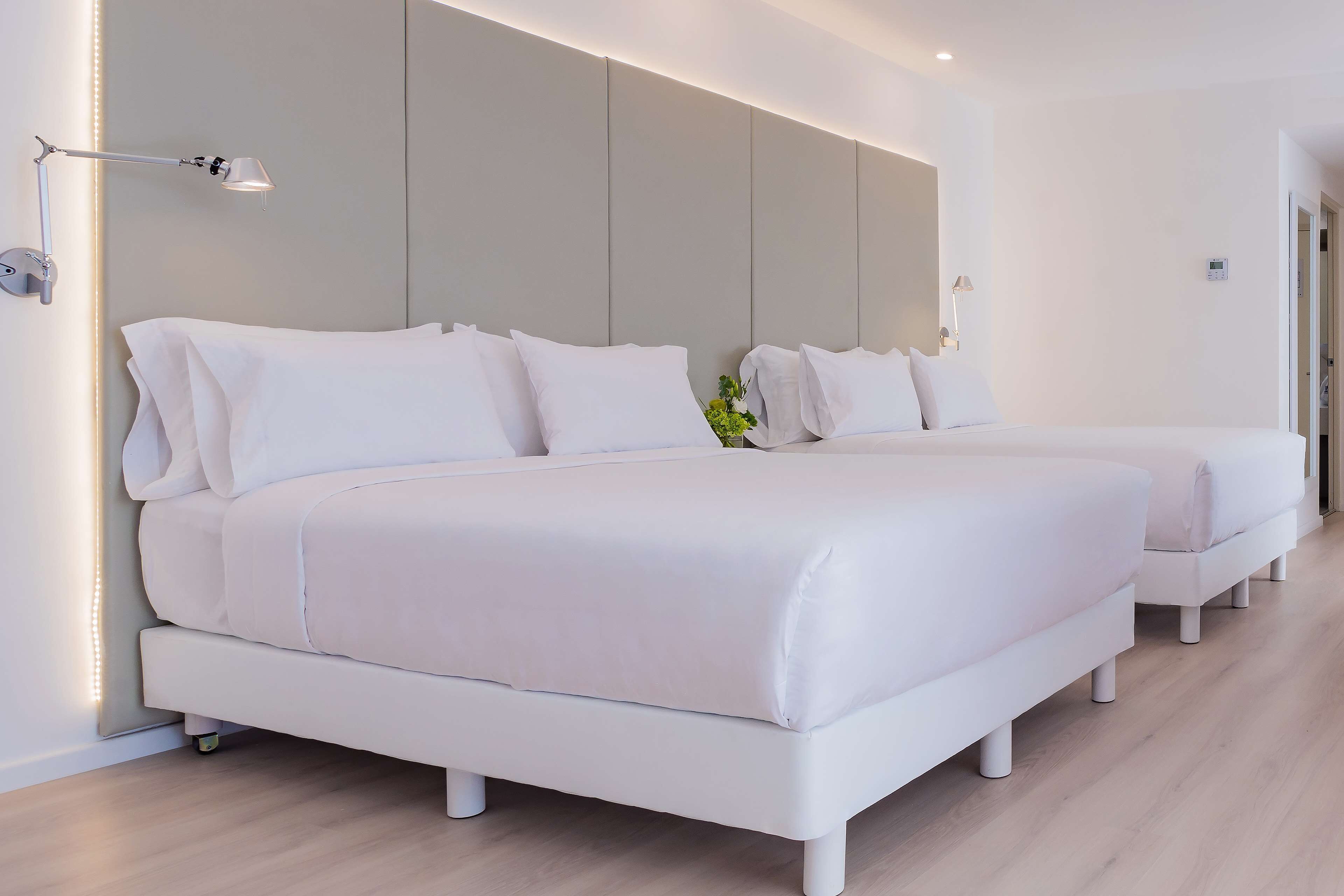 A modern hotel room with two white beds, a bedside table with a lamp, and a large wall-mounted panel.