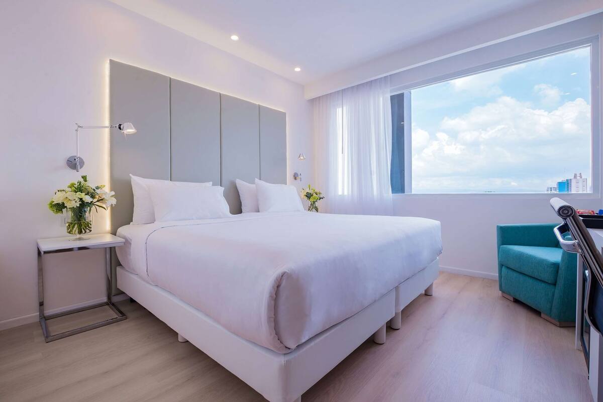 A modern hotel room with a large bed, a desk, a chair, and a window with a city view.
