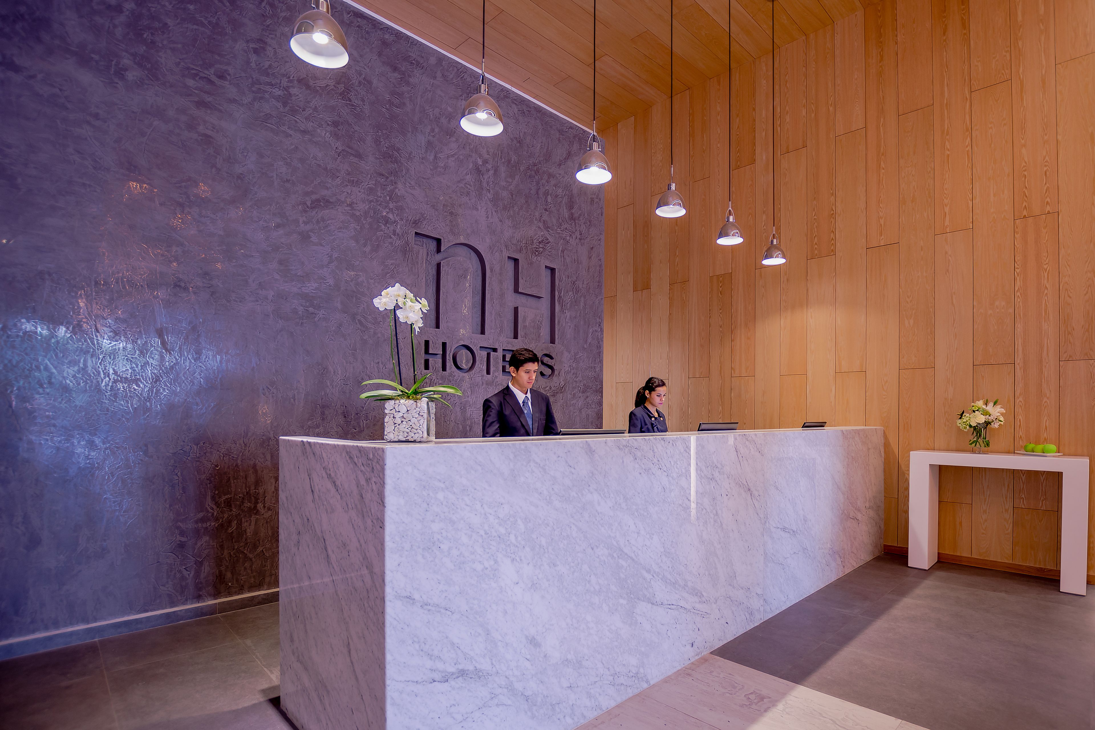 Una recepción con encimera de mármol, dos empleados atendiendo y un muro de madera con el nombre del hotel.