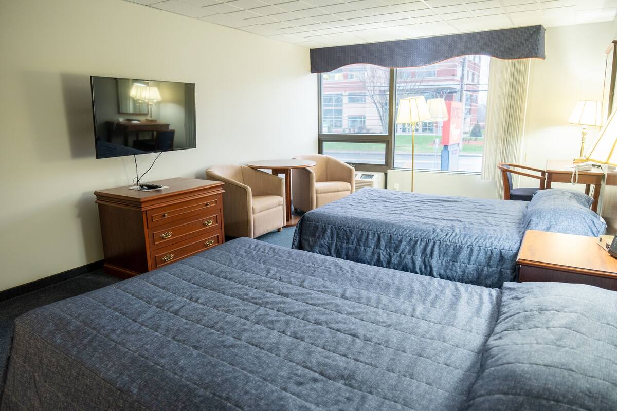 A hotel room with two beds, a TV, a desk, a chair, and a window with a view of buildings.