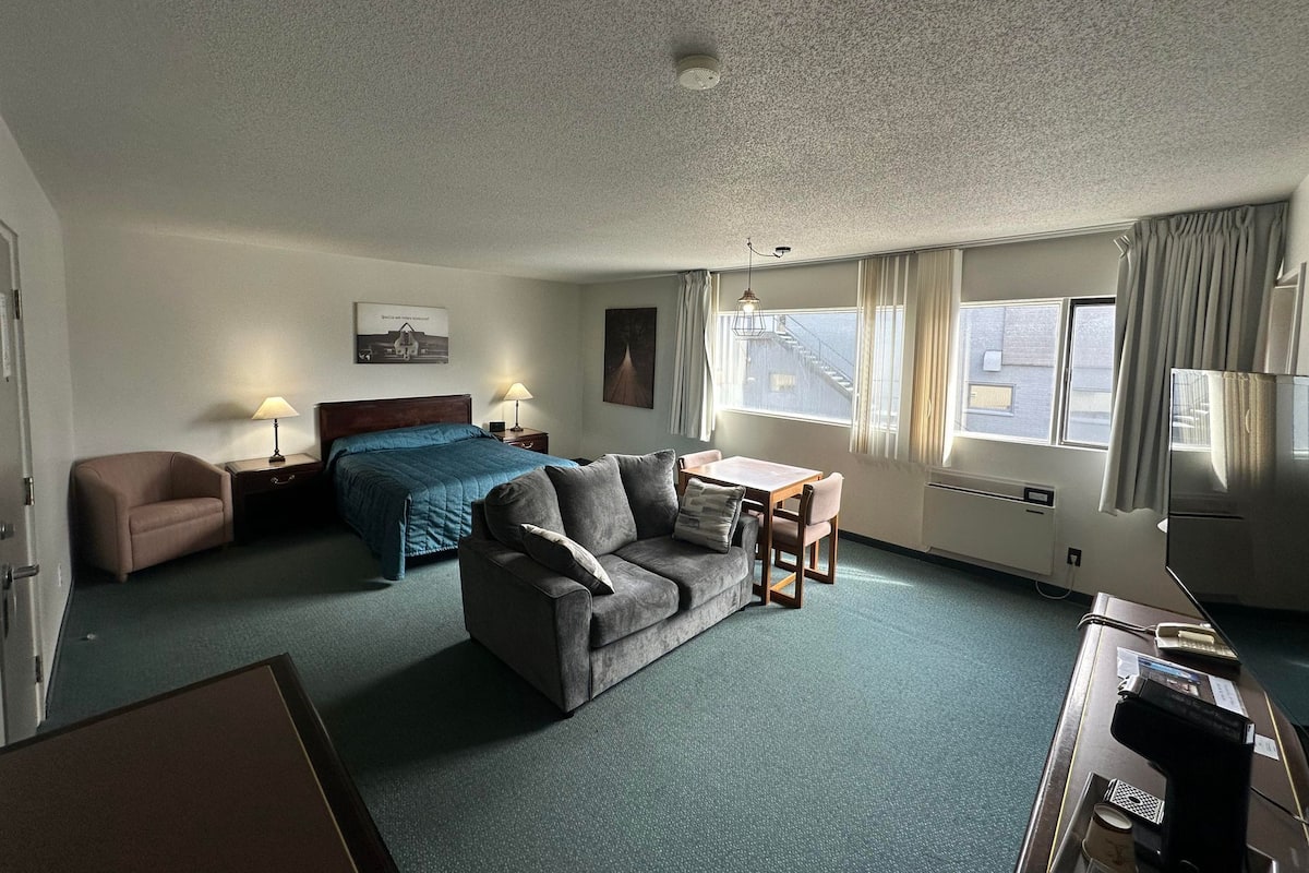 A hotel room with a bed, a sofa, a desk, and a chair. There is a window with curtains, a lamp, and a picture on the wall.