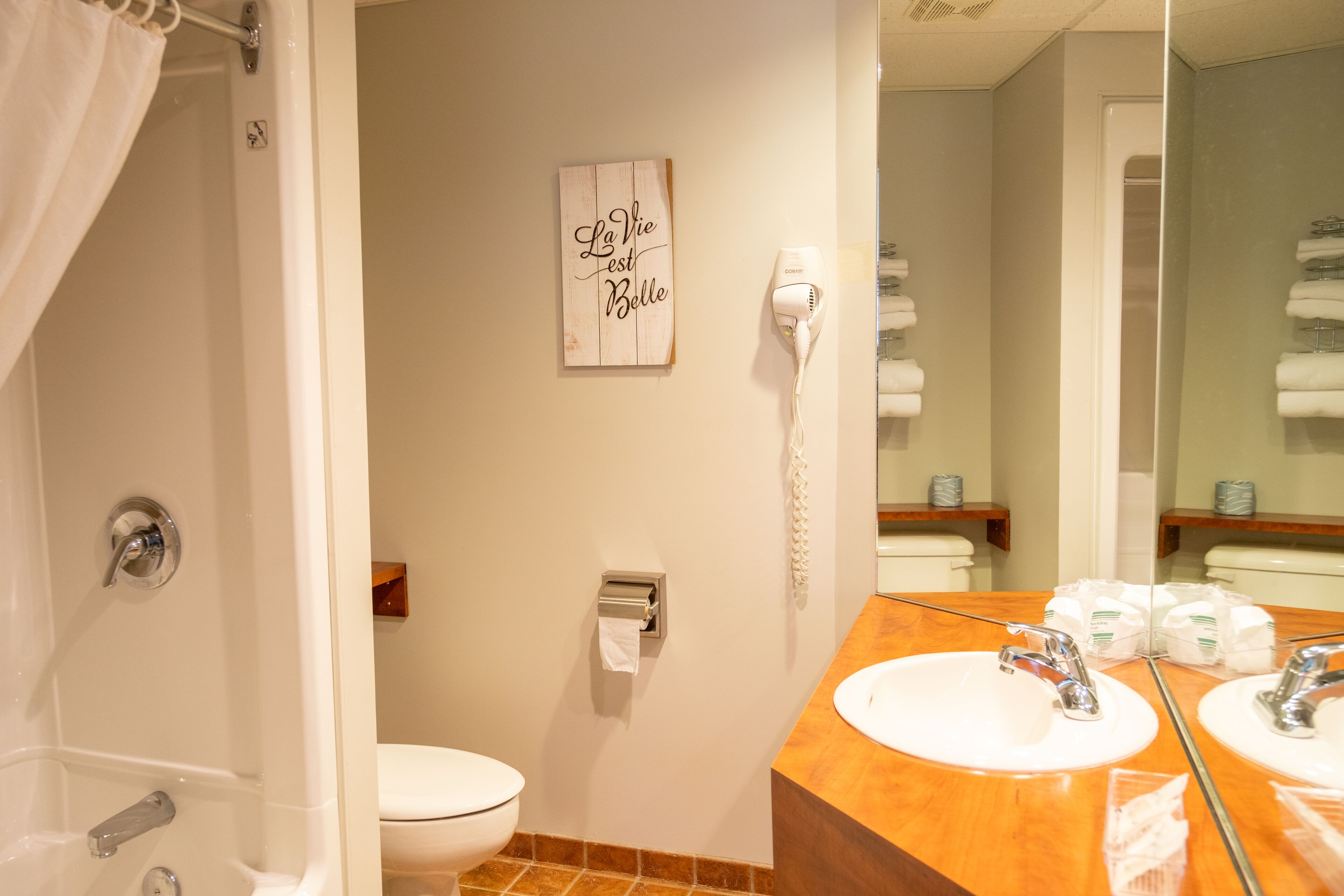 Une salle de bain avec des toilettes blanches, un panneau en bois sur lequel est écrit «La vie est belle», un sèche-cheveux et un miroir avec un porte-serviettes.