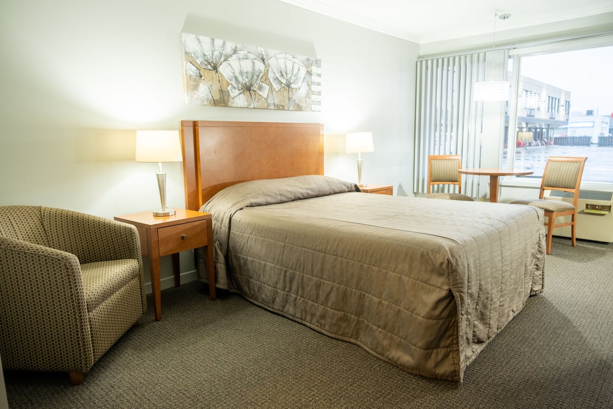 A hotel room with a large bed, two bedside tables, a chair, a small table, and a window with curtains.