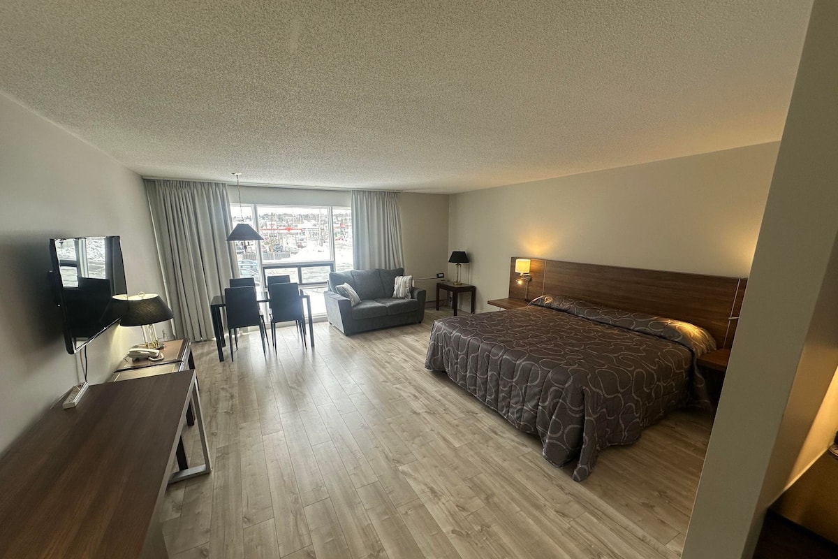A hotel room with a large bed, a sofa, a desk with a lamp, a TV, and a view of the city through the window.