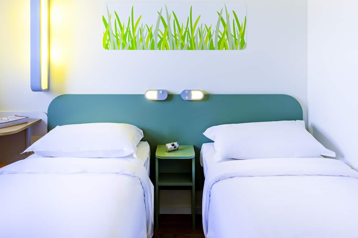 Ein Hotelzimmer mit zwei Einzelbetten, einem kleinen Nachttisch mit Lampe, einer Wandleuchte und einem Gemälde von Gras.