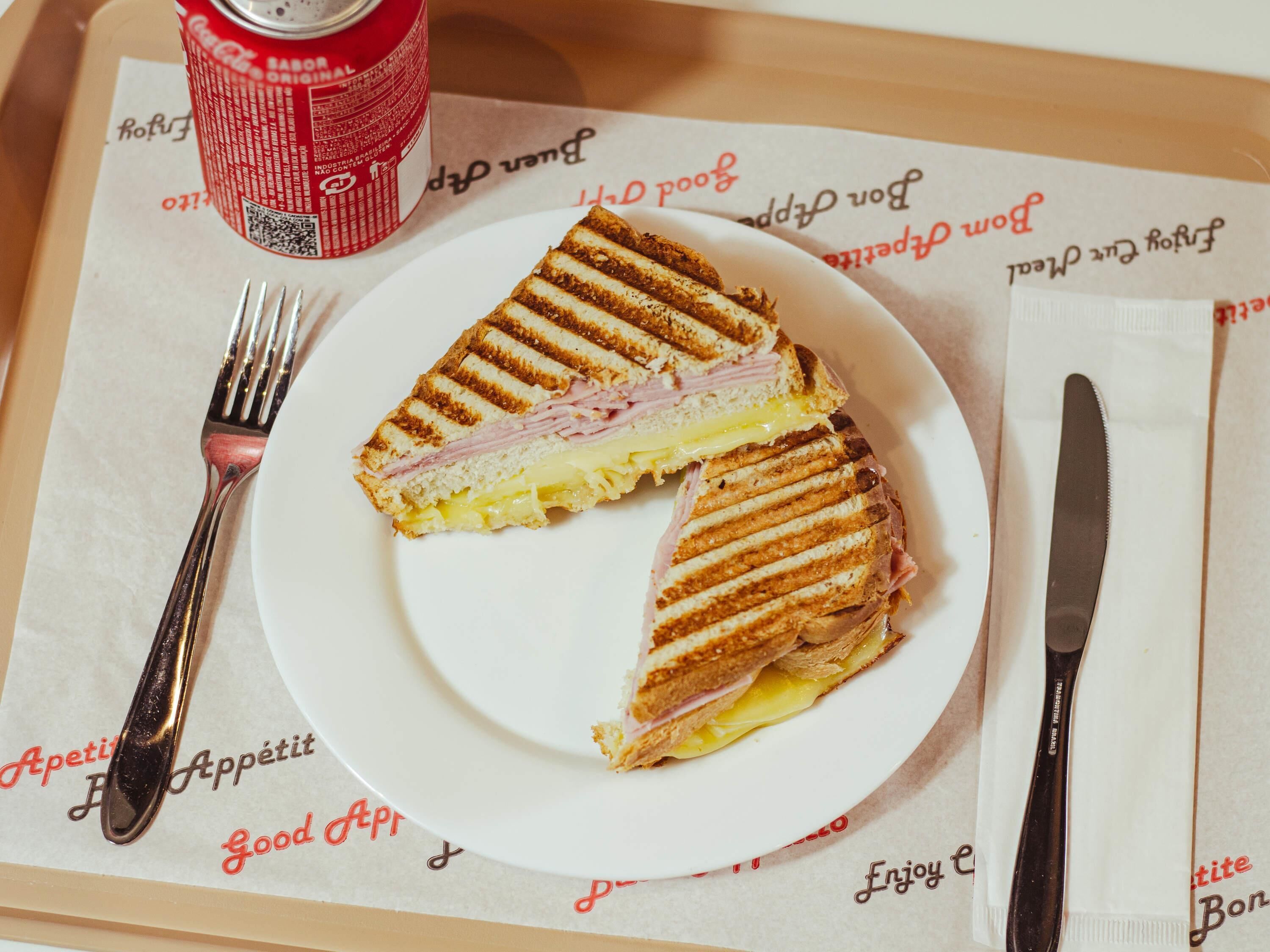 A plate with a grilled sandwich, a fork, a knife, and a red can on a tray with printed text.