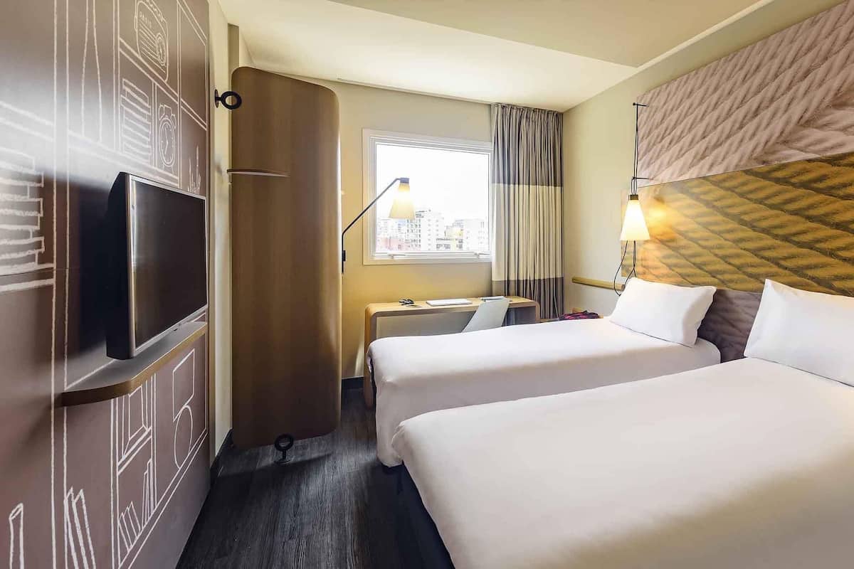 Une chambre d’hôtel avec deux lits, un bureau, une télévision et une fenêtre avec des rideaux.