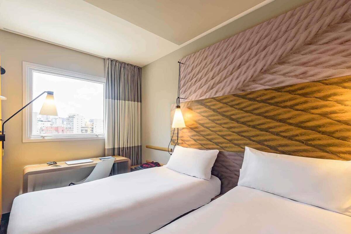 Ein Hotelzimmer mit zwei Betten, einem Schreibtisch, einem Stuhl, einer Lampe und einem Fenster mit Vorhängen.