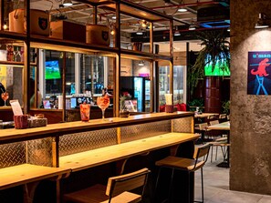 A restaurant interior with wooden tables and chairs, a bar area with drinks, and a large window with a view of the city.