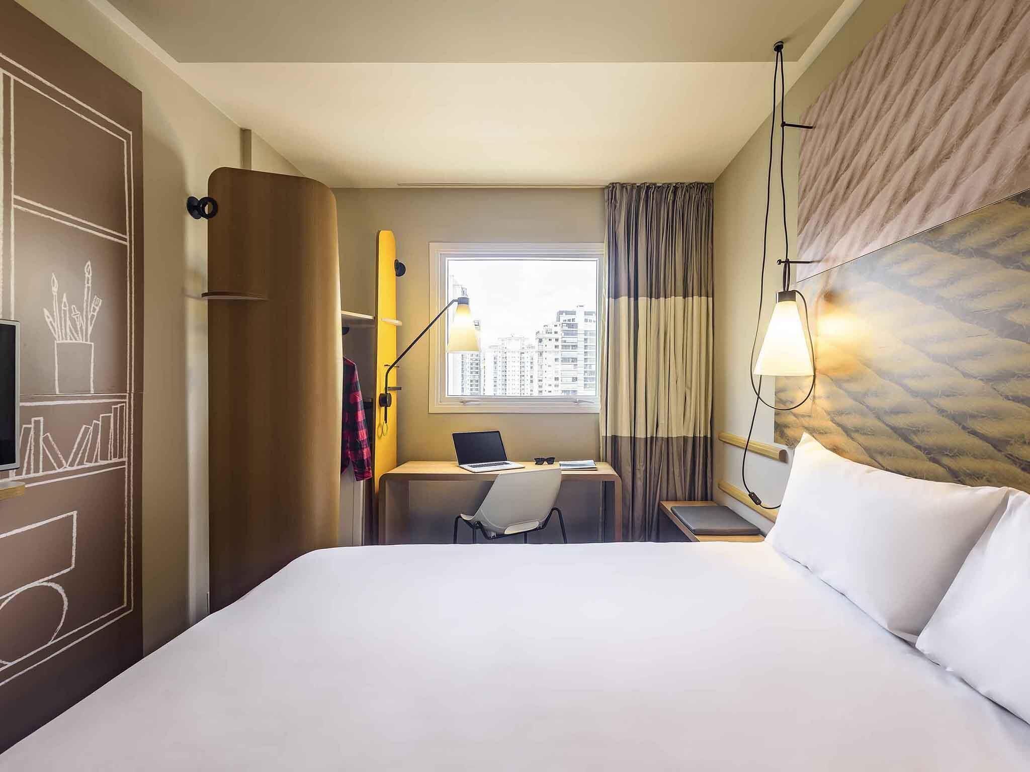 Une chambre d’hôtel avec un lit, un bureau avec un ordinateur portable, une chaise, une fenêtre avec des rideaux et une vue sur la ville.