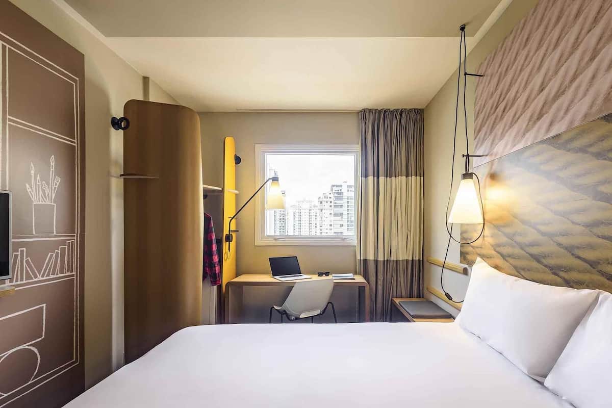 Ein Hotelzimmer mit einem Bett, einem Schreibtisch mit Laptop, einem Sessel und einem Fenster mit Vorhängen, mit Blick auf die Stadt.