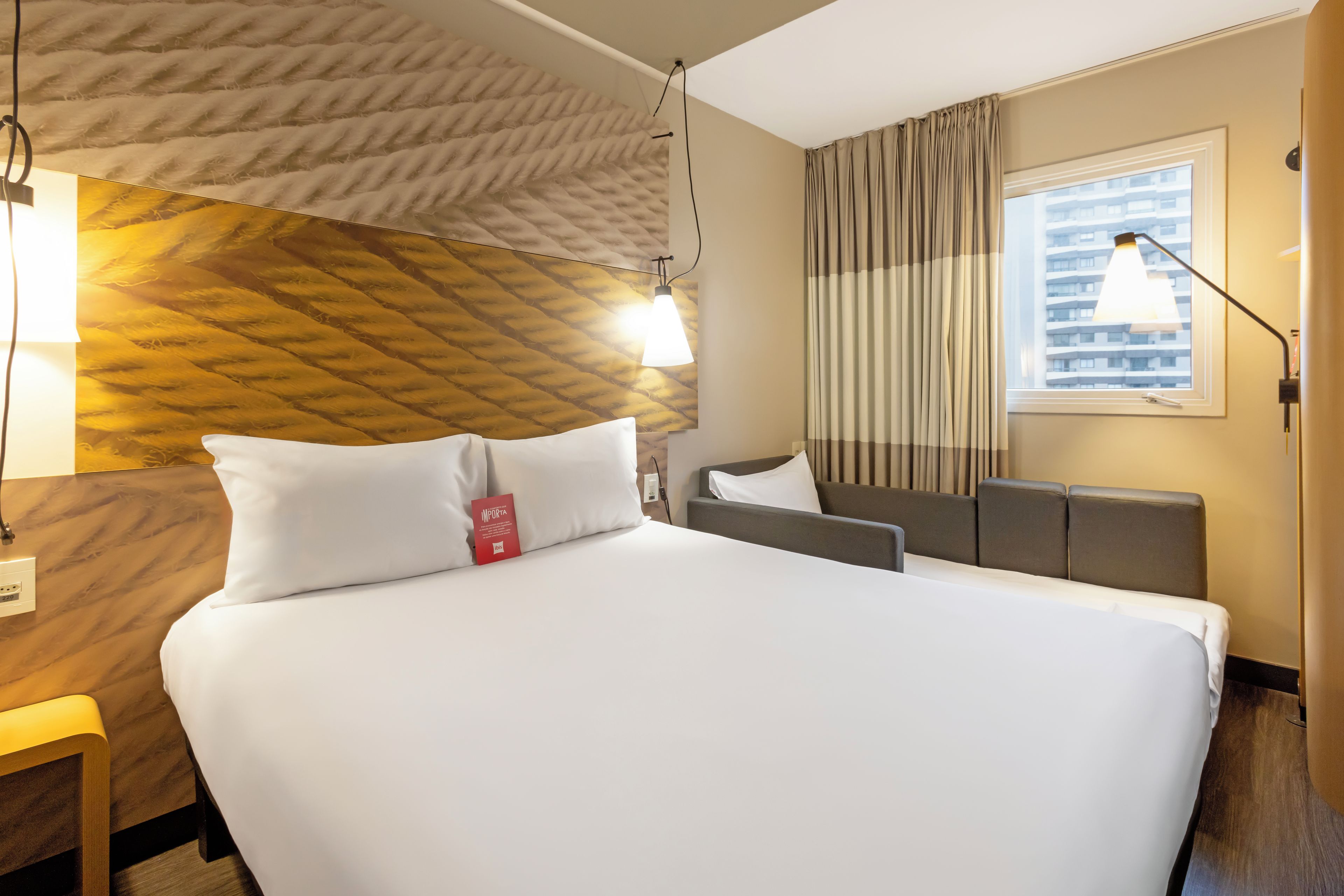 A hotel room with a bed, bedside table, lamp, and a window with curtains.