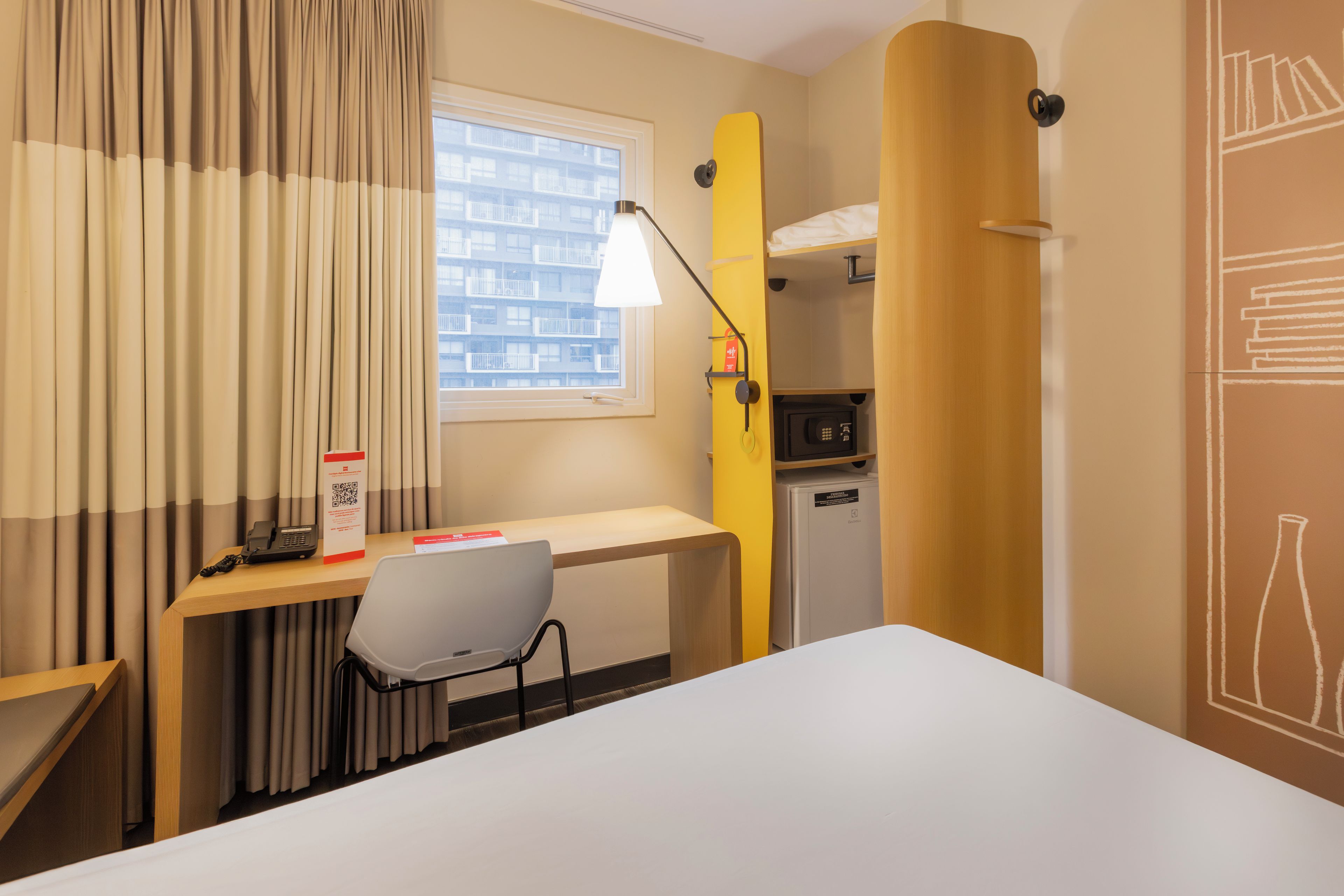 A hotel room with a desk, chair, bed, and a small kitchenette.
