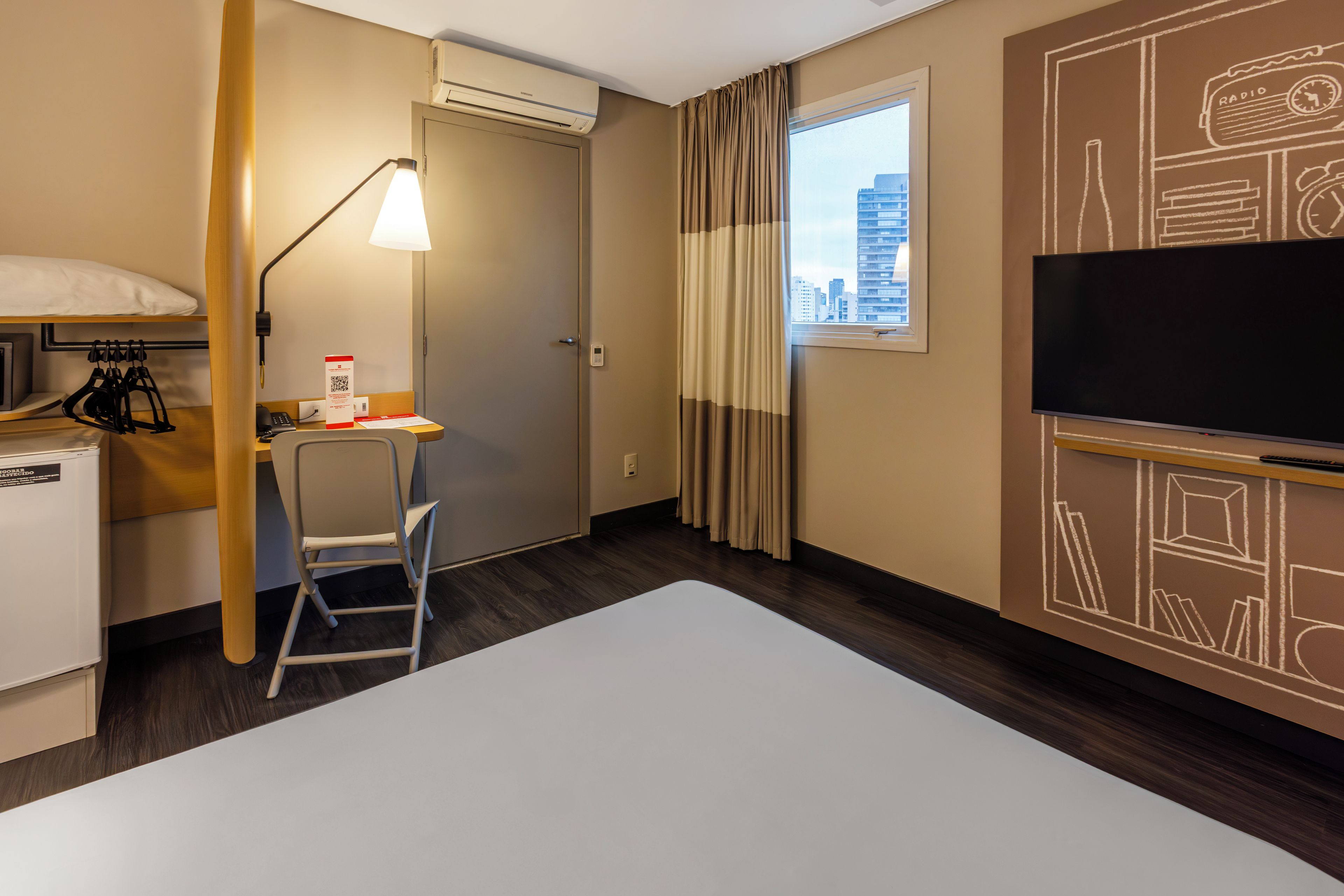 A hotel room with a desk, chair, bed, air conditioning unit, window with curtains, and a television.
