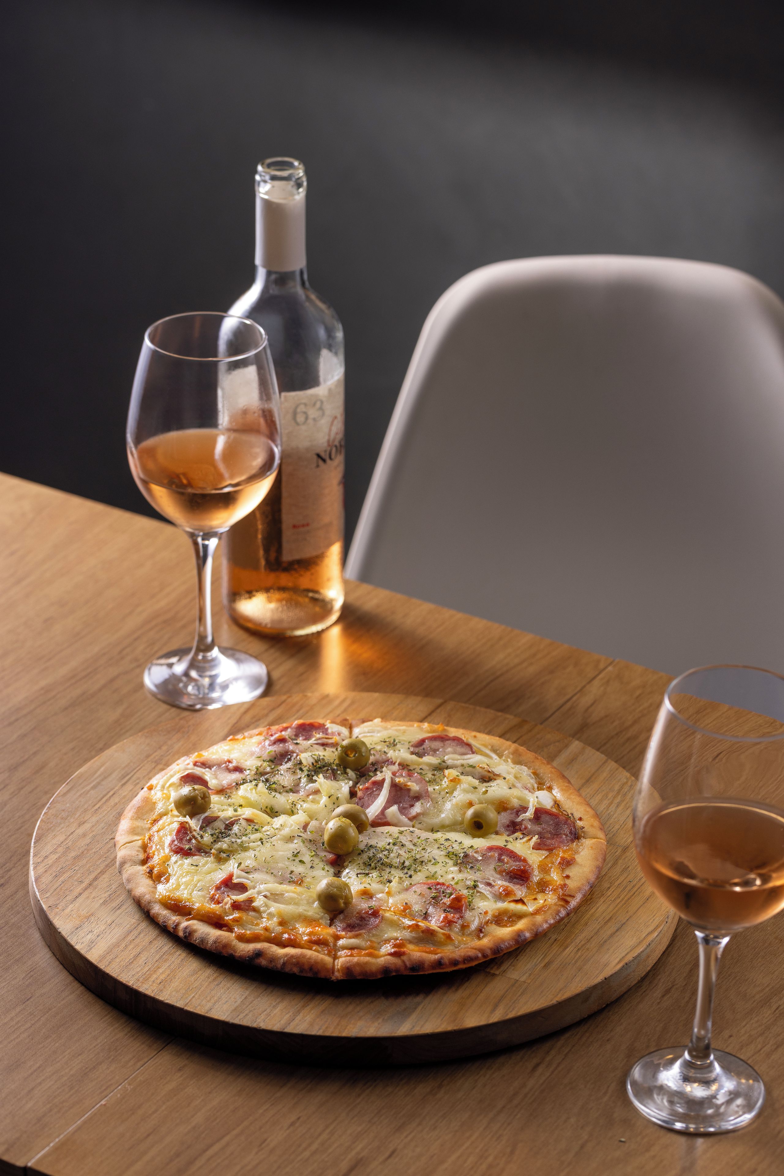 Une pizza sur une planche en bois, une bouteille de vin et deux verres à vin sur une table.