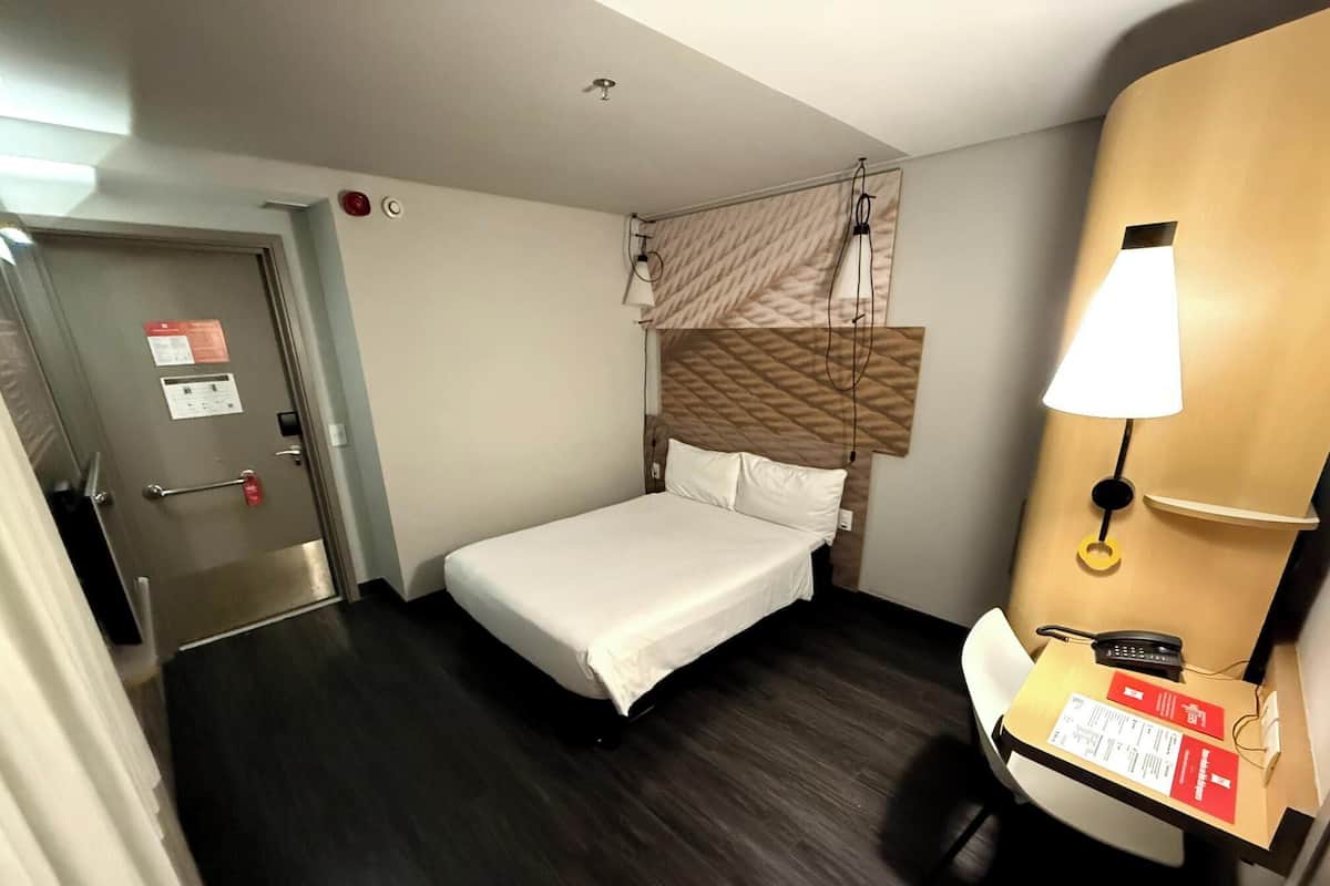 Une chambre d’hôtel avec un lit, une table de chevet avec une lampe, un téléviseur fixé au mur et une porte dotée d’une poignée et d’un bouton rouge d’appel d’urgence.