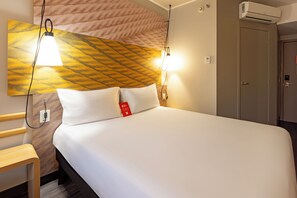 Une chambre d’hôtel comprenant un lit, une table de chevet, une lampe fixée au mur, un appareil de climatisation et une porte.