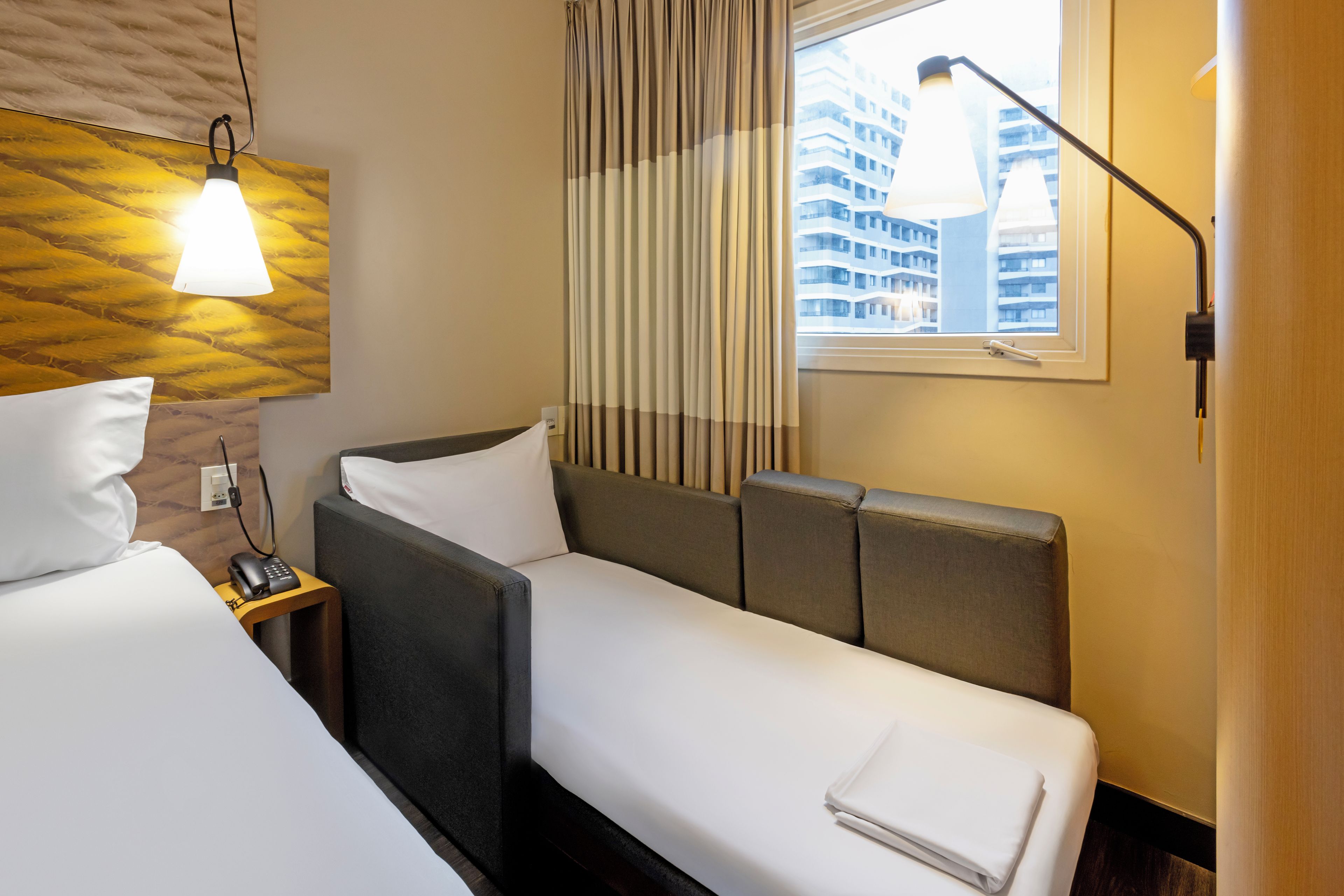 Habitación de hotel con una cama, un sofá, una mesita de noche con lámpara y una ventana con cortinas.