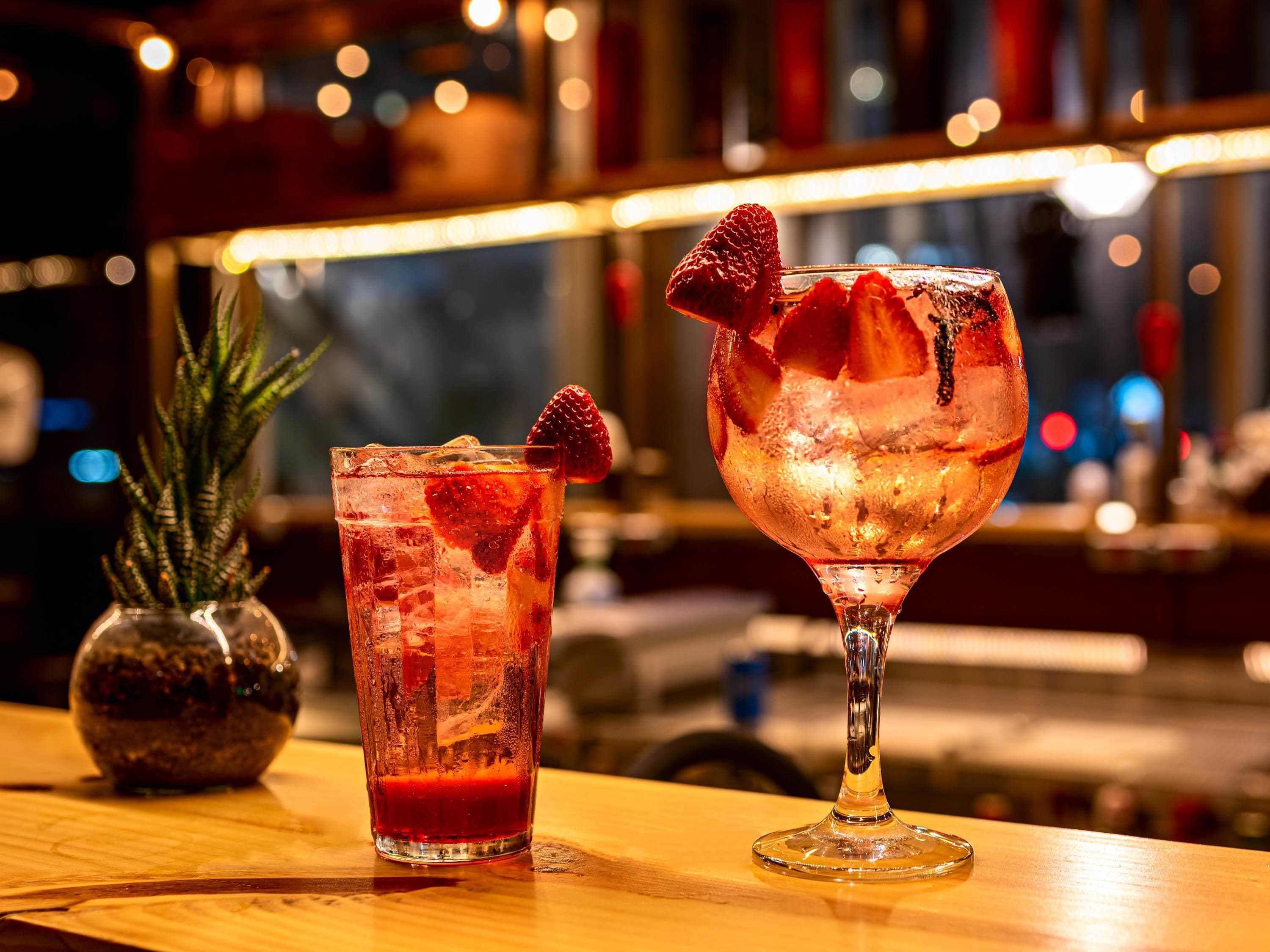 Two glasses of a red beverage with ice and a strawberry garnish on a wooden bar counter.
