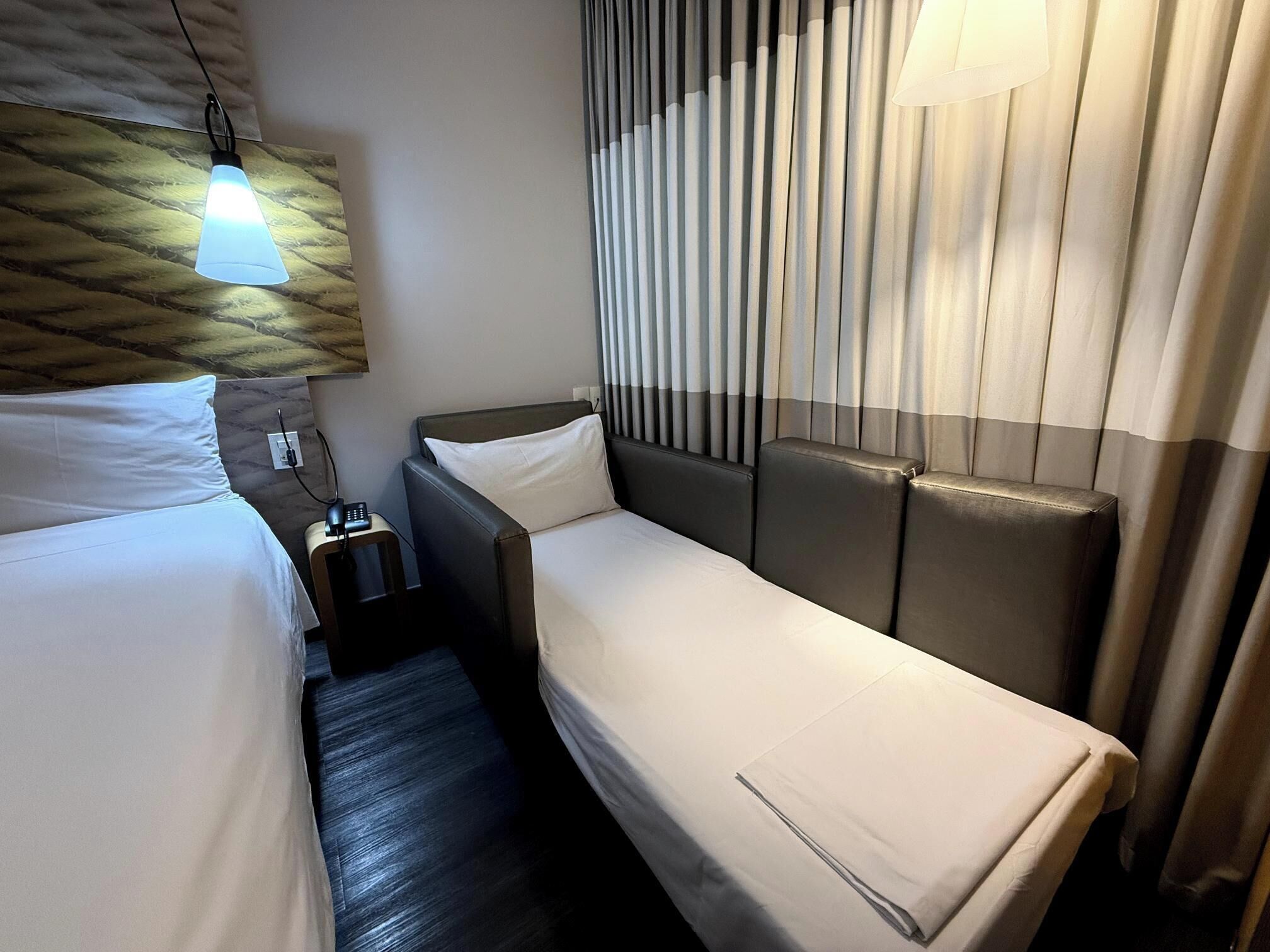 A hotel room with two beds, a nightstand with a phone, a wall-mounted light, and a window with curtains.