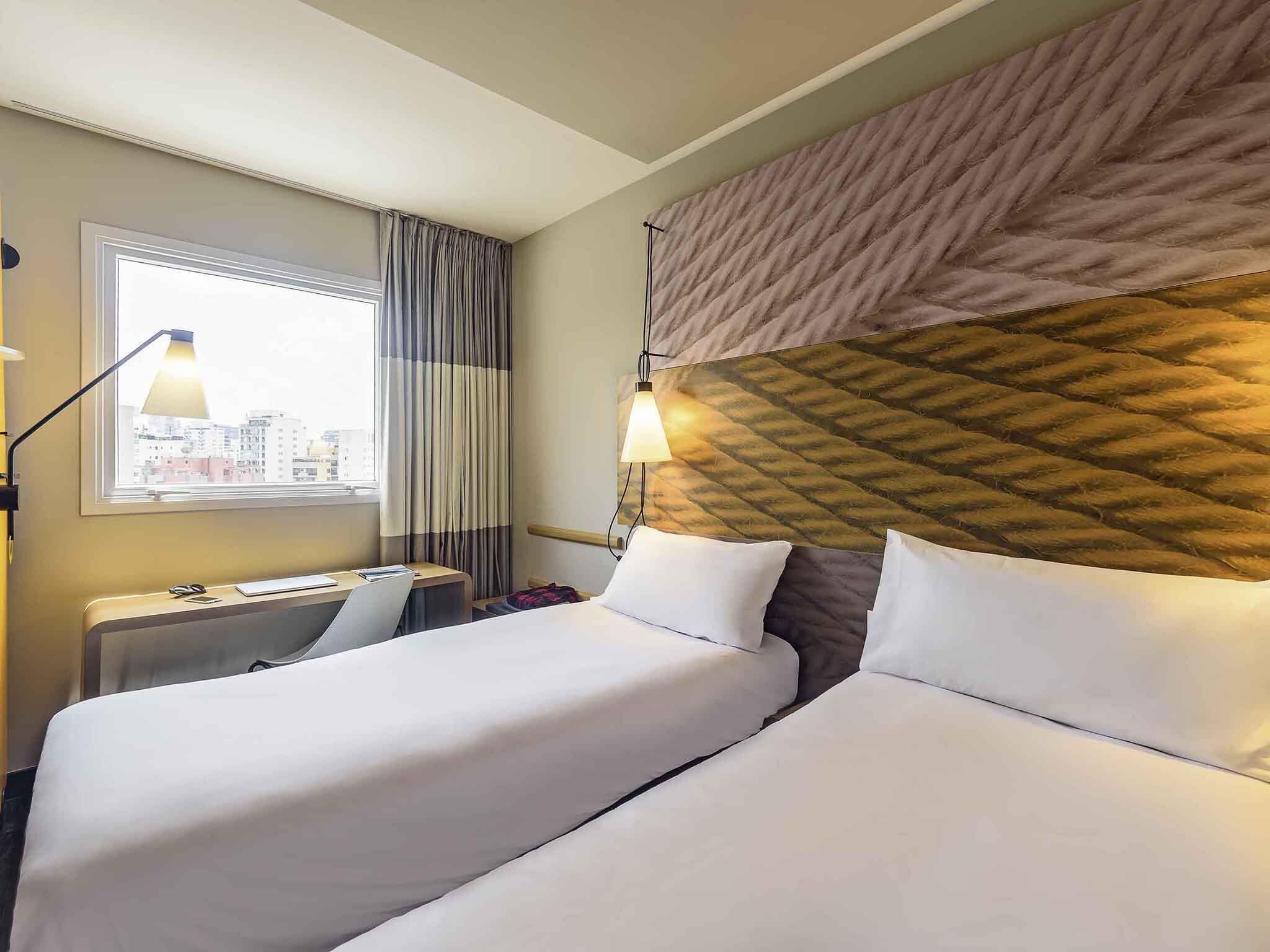Une chambre d’hôtel avec deux lits, une fenêtre avec des rideaux, un bureau avec une lampe et une vue sur la ville.