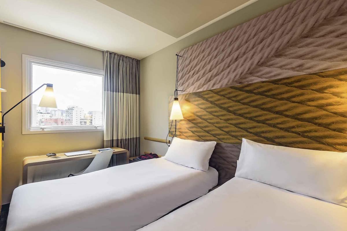 Ein Hotelzimmer mit zwei Betten, einem Fenster mit Vorhängen, einem Schreibtisch mit Lampe und Blick auf die Stadt.