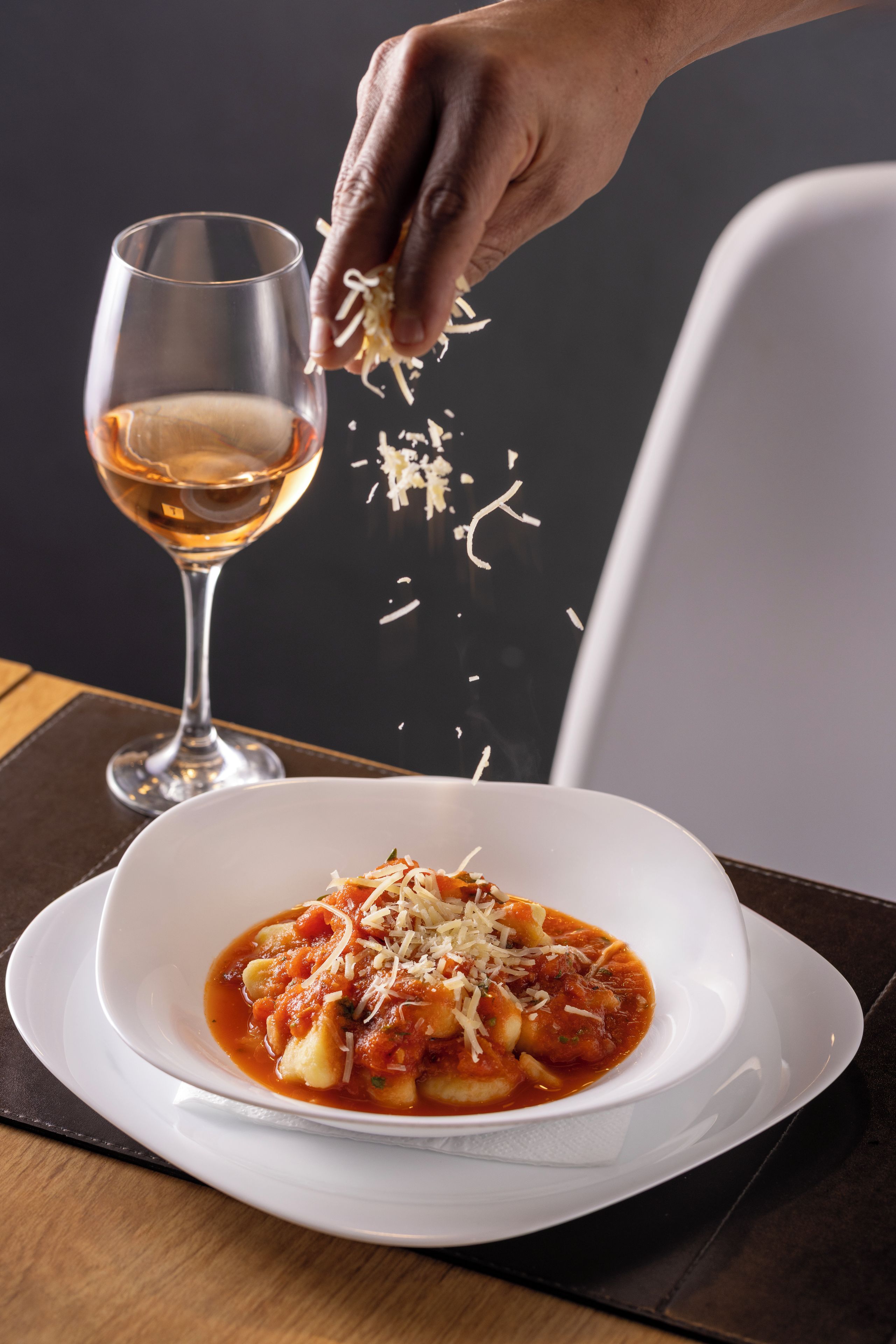 Un plat de pâtes nappées d’une sauce rouge et recouvert de fromage râpé, présenté sur une assiette blanche, et une main y ajoute du fromage supplémentaire.
