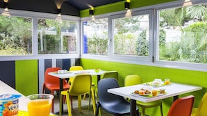 Une salle à manger aux couleurs vives, avec des tables et des chaises, une fenêtre donnant sur un paysage verdoyant, et un plateau contenant de la nourriture et des boissons.