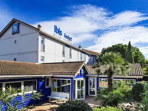 Un hôtel Ibis Budget à deux étages, doté d’un toit en tuiles, d’une entrée bleue et d’un jardin agrémenté de palmiers.