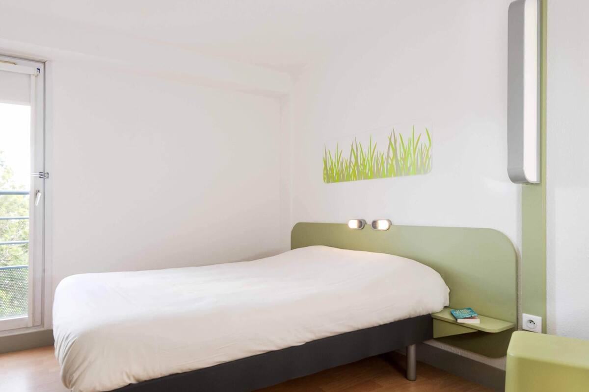 A bedroom with a bed, a nightstand, a wall-mounted lamp, and a window.