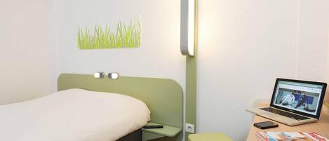 Habitación de hotel con una cama, un escritorio con una computadora portátil, una mesita de noche verde y una lámpara de pared.