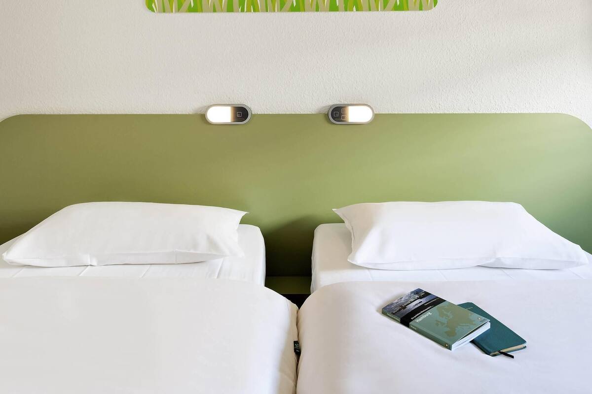 Ett hotellrum med två sängar, båda med vita sängkläder och kuddar. Ovanför sängarna finns en väggmonterad lampa, och en grön dekorpanel med mönster som liknar gräs.