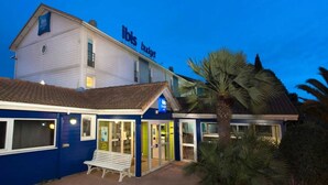 Un bâtiment à deux étages portant l’enseigne « ibis budget », avec une entrée principale dotée de portes vitrées, un banc à l’extérieur et un palmier à proximité.