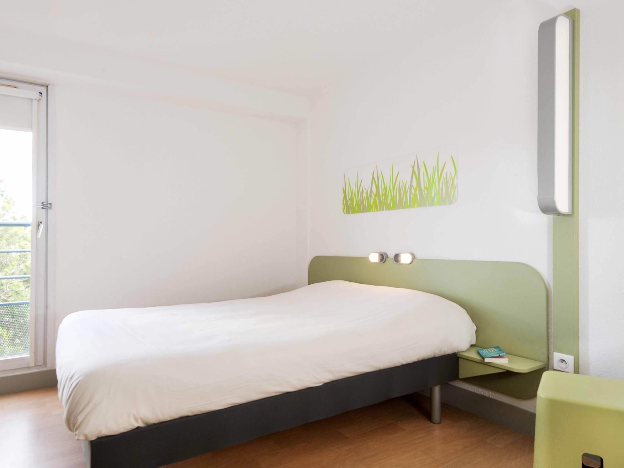 Une chambre à coucher avec un lit, une table de chevet, une lampe fixée au mur et une fenêtre.