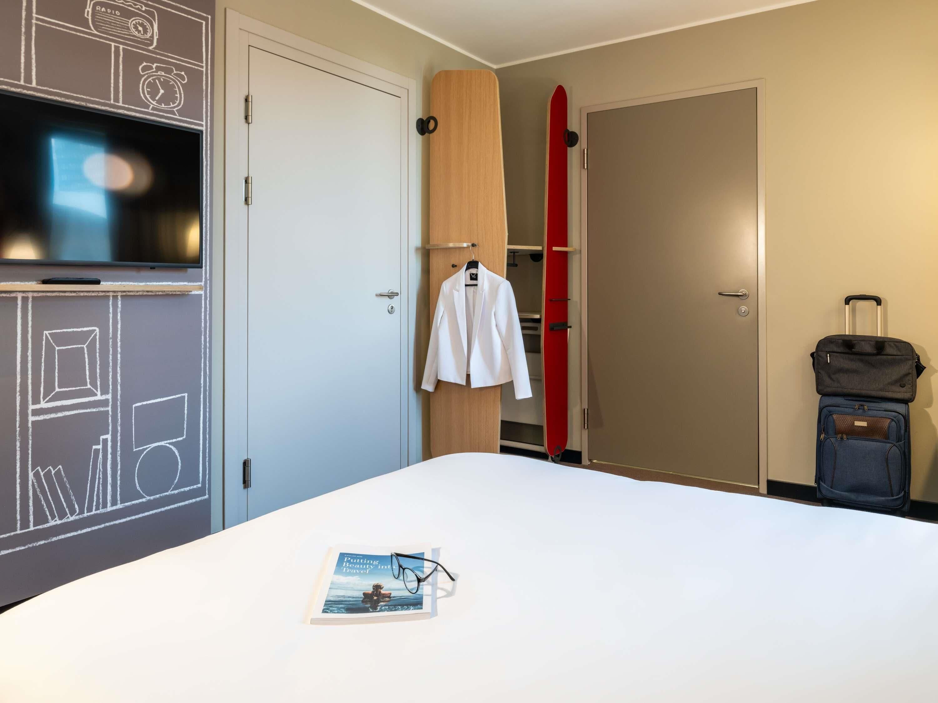 A hotel room with a bed, a white jacket hanging on a wooden closet door, a red surfboard leaning against the wall, and a suitcase on the floor.