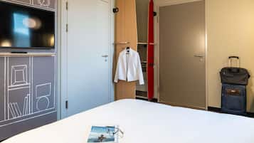 A hotel room with a bed, a white jacket hanging on a wooden closet door, a red surfboard leaning against the wall, and a suitcase on the floor.