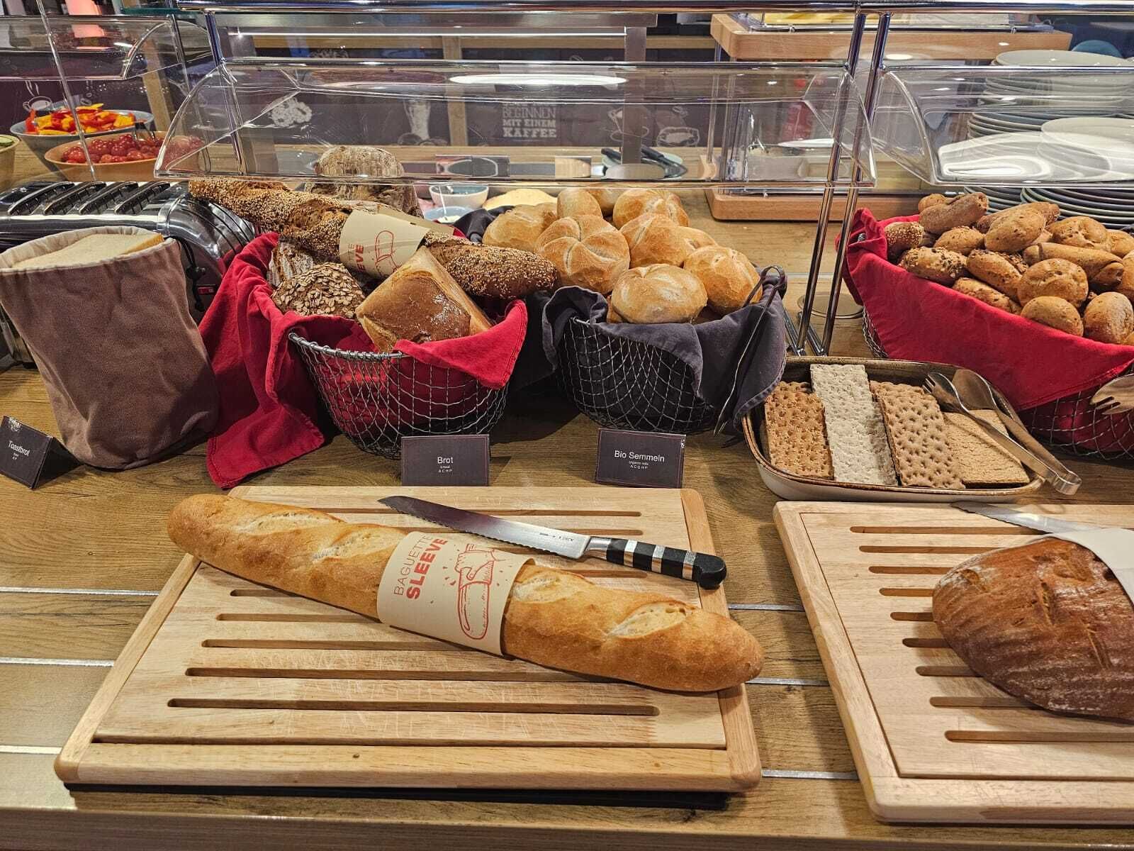 Une vitrine de boulangerie présentant diverses sortes de pains et de viennoiseries, notamment une baguette et un pain de mie, le tout disposé sur des planches à découper en bois.