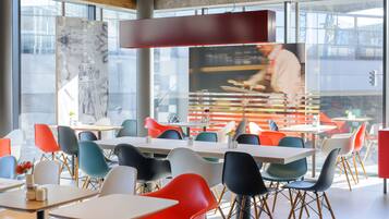 Un espace repas moderne avec des chaises et des tables aux couleurs vives, de grandes fenêtres et une cuisine ouverte.
