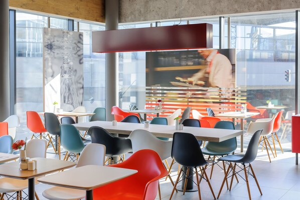 A modern dining area with colorful chairs and tables, large windows, and a visible kitchen area. - Ibis Wien Hauptbahnhof (Vienna)