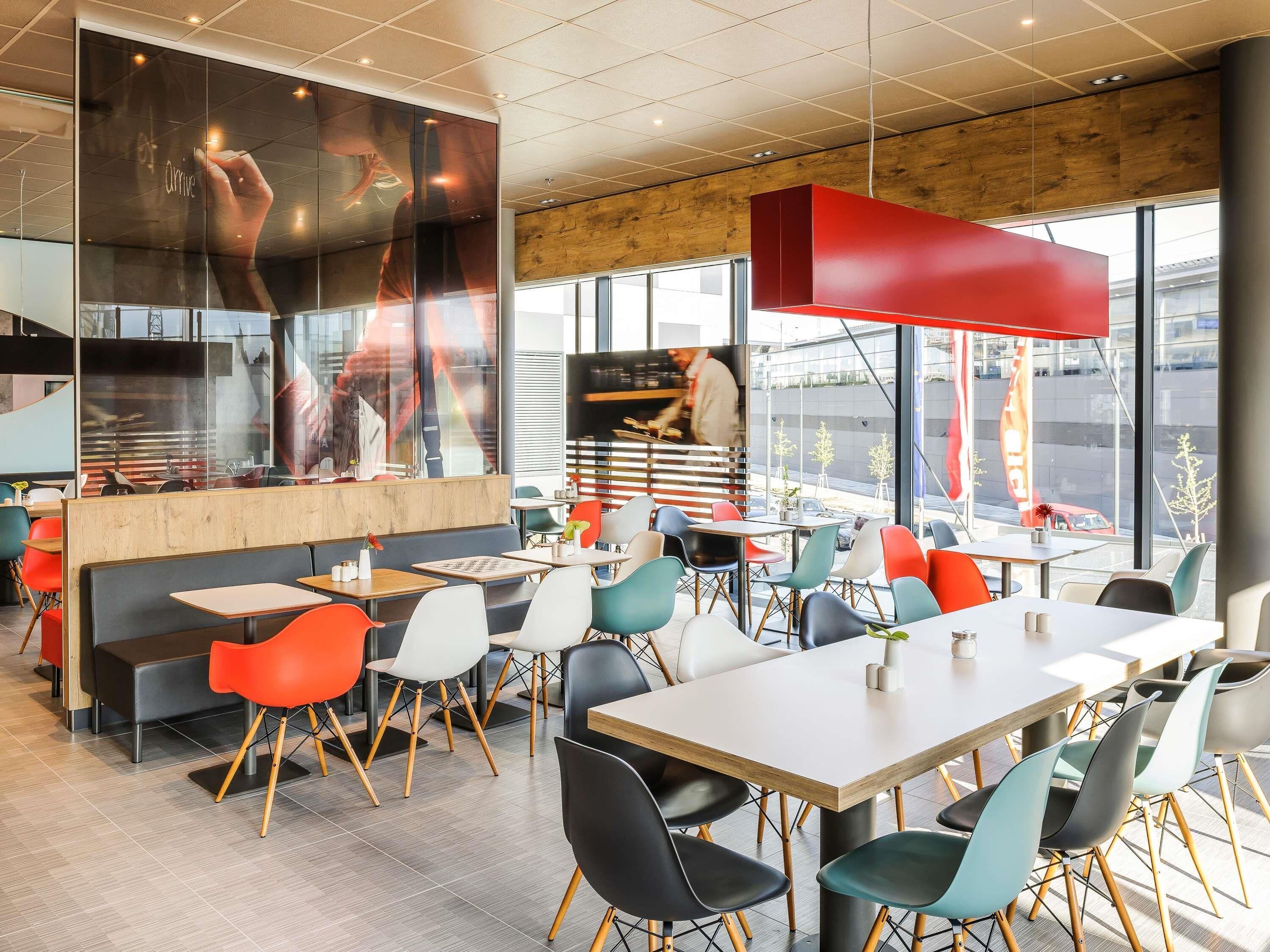 Ein modernes Restaurant-Interieur mit verschiedenen Sitzmöglichkeiten, darunter Sitzecken und Tische, und einem großen Fenster mit Blick auf die Straße.