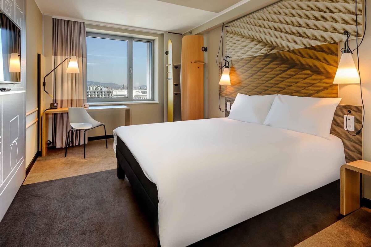 Une chambre d’hôtel dotée d’un grand lit, d’un bureau avec une chaise, d’une fenêtre offrant une vue agréable et d’une tête de lit au design original et à la texture particulière.
